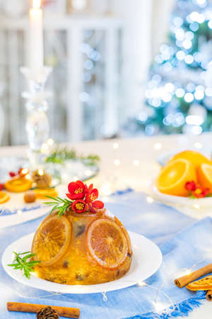 Traditional English Christmas pudding decorated with oranges and flowers in festive table setting with fruits, spices, lights of Christmas tree on background. Copy space. Magical mood. Vertical card.の写真素材