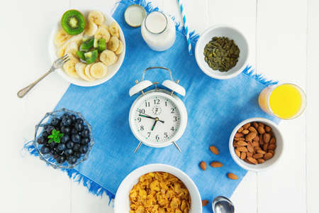 Time for breakfast.Top view alarm clock with healthy morning food. Bowl with cereals, nuts and berries, milk, fresh juice on blue napkin and white wooden table. Day diet planning and healthy eating.の写真素材