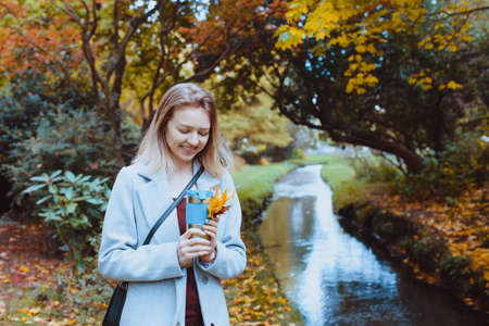Smiling beautiful young woman in coat enjoying hot drink in her reusable thermos cup and walking in the autumn park near the river. Romantic fall mood. Vintage toning. Selective focus. Copy space.の写真素材
