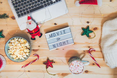 Top view Christmas Movie night concept. Flat lay composition with Movie night message on the board, laptop, popcorn bowl, decor, a cup of cocoa with marshmallows and warm plaid on wooden backgroundの写真素材