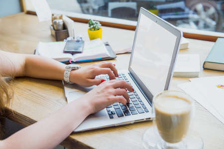 Close up a woman working on a laptop and drinking a cup of coffee while sitting in a cafe or co-working with street view. Soft selective focus. White blank screen. Copy space, Selective focus.の写真素材