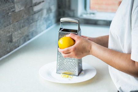 Female hands grating fresh whole lemon on metal hand grater, making zest on her kitchen table....