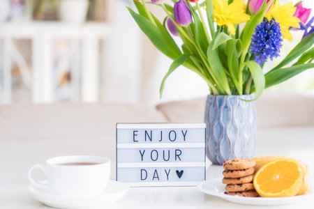 Good morning concept. Romantic breakfast - fresh flowers in vase, cup of hot drink, cookies, orange, lightbox with message Enjoy your day on marble table with light interior view. Close up. Copy spaceの写真素材