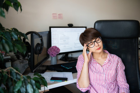 Feel tired or depressed woman in glasses and striped shirt sitting at her desk at home workspace...