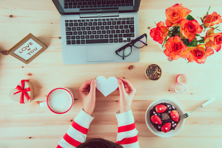 Top view woman holding blank white heart shape greeting card above her working place with a laptop. Surprise idea on Valentines Day. Sweets, a cup of coffee, flowers, and gift box on the wooden desk.の写真素材