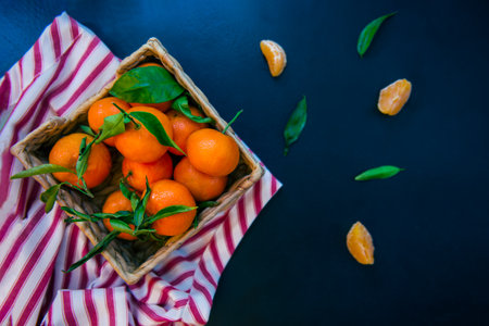 Top view Fresh ripe mandarins, clementine, tangerine with green leaves in the straw bowl on a striped napkin and dark blue stone background. Local citrus fruit harvest. Selective focus, flat lay.の写真素材