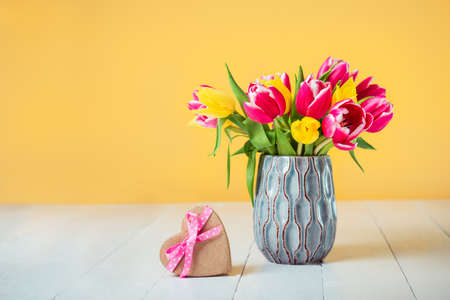 Craft heart shape gift box with satin ribbon bow and spring bouquet of pink and yellow tulip flowers in a vase on the white wooden table and light yellow background. Greeting card mockup. Copy sapce.の写真素材