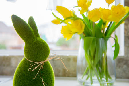Cute green grass bunny rabbit and bouquet of fresh spring yellow tulips and daffodils flowers on the...