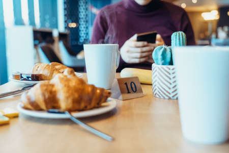 Steaming hot tea cup and fresh croissants on the table where young man using his phone during his breakfast in the hotel restaurant. Selective focus, copy space.の写真素材