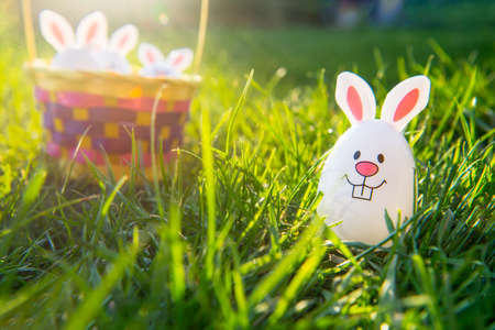 Creative Easter composition of Funny Easter egg with bunny ears on green grass and straw basket with other eggs inside. Sunny festive mood. Fairy tale Easter hunt. Selective focus. Copy space.の写真素材
