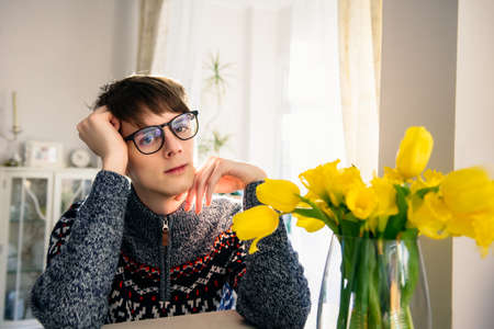 Portrait of young sensual teenager man seating at the table with yellow flowers bouquet on light interior background. Fight with gender stereotypes. Equality concept. Selectivr focus, copy space.の写真素材