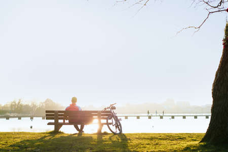 Back view man sitting on the bench at the bank of parks lake and enjoying the sunset. Enjoy nature, healthy break, digital detox. Selective focus, copy space.の写真素材