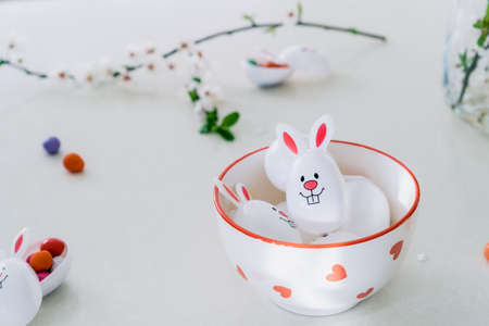 Funny Easter eggs containers filled with candy and chocolate eggs with bunny ears in a bowl on the white kitchen table. Festive spring Easter composition. Selective focus, copy space.の写真素材