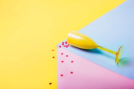 Red Heart-shaped confetti falling from yellow wine glass lying on the pink and blue podium on the yellow background. Creative love concept. Valentines Day. Trendy, minimalist card with copy space.の写真素材