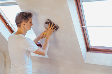 A young man scratching with a spatula and removing old wallpaper on a wall of a room. Do it yourself...