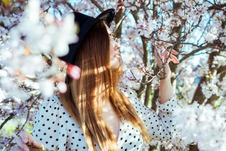 Young brunette woman in hat and dress near a white blooming tree. Generation Z girl enjoy spring mood during sunny day. Selective focus, copy spaceの写真素材