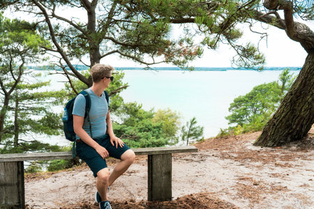 Man with backpack sitting on the bench and enjoying sea view during hiking walk in summer forest....