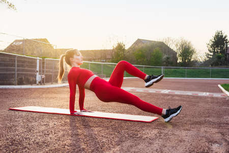 Young fitness woman in red sportswear doing reverse plank knee tuck exercise on the mat. Outdoors...