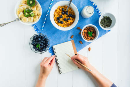 Morning habits of successful people. Day planning and healthy meal. Woman writing in notebook on the served for healthy vegetarian breakfast white wooden table. Top view, Selective focusの写真素材