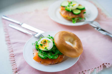 Fresh bagel sandwiches with salmon, soft egg, salad, microgreen sprouts, cucumber and cream cheese served on pink napkin with cutlery on the white kitchen table. Healthy Breakfast. Selective focusの写真素材