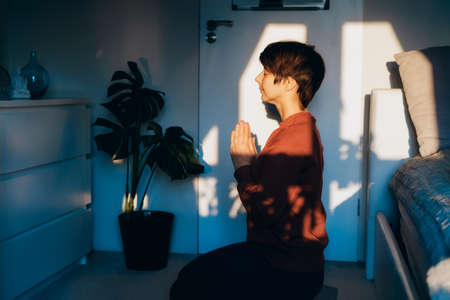 Woman with closed eyes sitting on a mat in the bedroom, doing Namaste in the sunset light. Praying ritual with faith to buddha statue at home. Zen, inner peace, grateful practice. Personal fulfillmentの写真素材