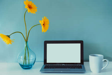 The modern workplace - desk with laptop mockup white empty screen, coffee cup, and fresh yellow gerbera flowers in the vase on the blue background. Workspace in home office. Color in interior design.の写真素材
