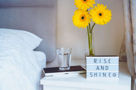 Good morning concept. Inspiration Motivational Life Quotes "Rise and shine" message on a lightbox with notebook and glass of water and fresh sunny yellow gerbera flowers on the bedside table.の写真素材