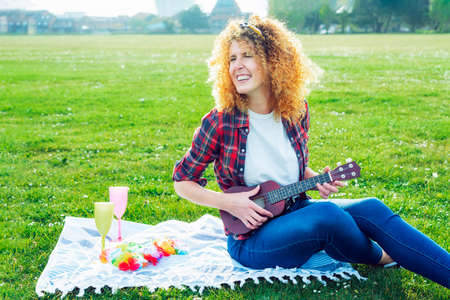 Happy laughing curly hair woman playing ukulele guitar while sitting on lush green grass in a city park at a picnic. Summer positive mood. Having fun with friends offline. Enjoy the moment.の写真素材