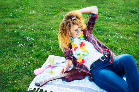 Happy laughing curly hair woman relaxing after playing ukulele guitar on lush green grass in a city park at a picnic. Summer positive mood. Having fun with friends outdoors offline. Enjoy the moment.の写真素材