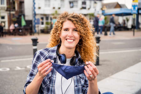 Young happy caucasian woman takes off a protective face-covering mask outdoors against European city...