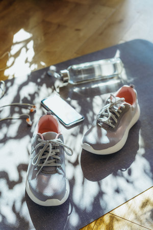 Bottle with water, smartphone, earphones and sneakers on the mat background with direct sunlight and shadows. Set for running and sports activities . Fitness, healthy and active lifestyle. Vertical.の写真素材