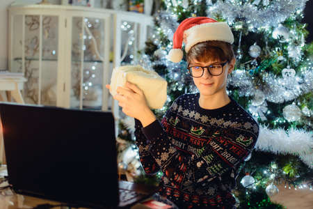 Young man with a red Santa hat holding gift box during online family greeting at home at Christmas...
