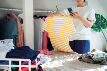 Woman taking photo of t-shirt using her smartphone for selling or donating her clothes. Decluttering...