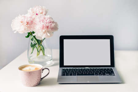 Light cozy workplace. White office table desk with a cup of fresh coffee, open laptop with blank screen and vase with blooming peonies. Minimalist workspace. Freelance, online learning, home office.の写真素材