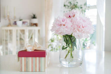 Fresh bouquet of tender pink peonies flowers and gift box with satin bow on white table with a light classic interior design background. Festive flowers as gift. Mockup for greeting card design.の写真素材