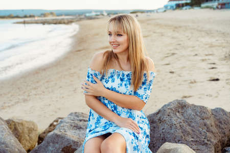 Young blonde woman in summer dress sitting on the rocks on the beach and enjoying the moment. Caucasian girl relaxing and enjoying peace on vacation. Selective focus, copy space.の写真素材