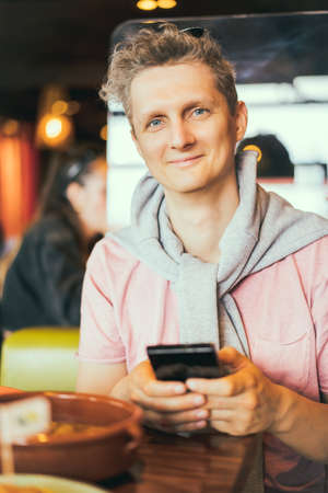 Portrait of a smiling young man using mobile phone while sitting in cafe. Make food photo for social networks, blogging, chatting, watching the video. Selective focus.の写真素材