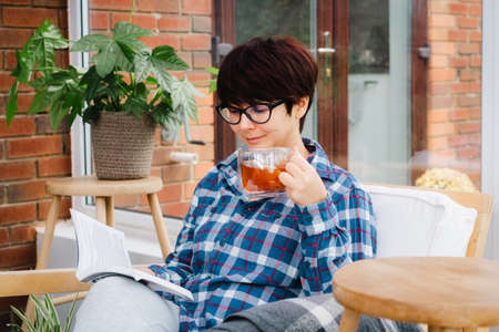 Woman in the glasses sit on the armchair and relax while reading a book and drinking hot tea at home...