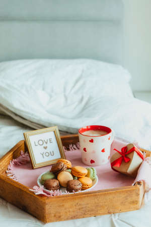 Valentines day breakfast in bed for Lover. Love you card in the frame, a cup of coffee or cocoa, macaroons, heart-shaped gift box with red ribbon on the wooden tray. Romantic morning. Copy spaceの写真素材