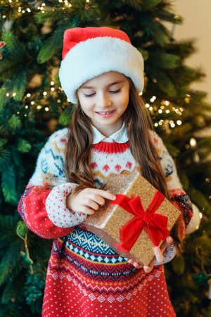 A happy smiling little girl in Santa hat and festive knitted sweater standing with gift box near the...
