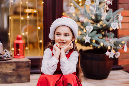 Smiling cute preschool kid girl in santa hat and red dress sitting on the porch of a house with decorated Christmas tree. Waiting for Santa and christmas holidays. Selective focus.の写真素材