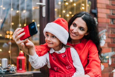 Smiling mother with daughter in Santa hat making selfie via phone while sitting on house porch with...