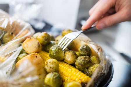 Male hands checking baked brussels sprouts, parsley root, corn plant based dish. Mixed vegetables in a baking bag on the tray. Cooking healthy vegan lunch. Vegetarian cristmas dinner. Selective focusの写真素材