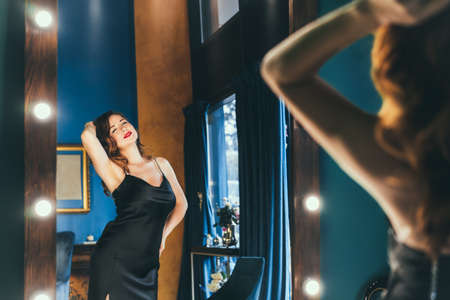Sexy young woman in elegant cocktail silk black dress dancing and posing in mirror reflection in luxury interior. Fashionable and self-confident lady. Celebrity, superstar lifestyle. Selective focusの写真素材