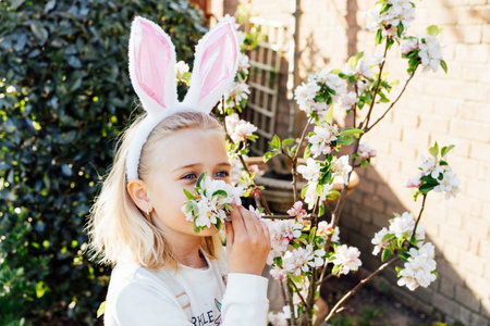 Adorable little blonde girl in bunny ears costume sniffing blooming tree branch outdoors on a spring...