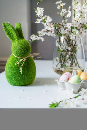 Green easter bunny with blooming flowers on tree branch in the vase and colored easter eggs in container on the white table. Happy Easter spring vertical card. Selective focus. Copy space.の写真素材