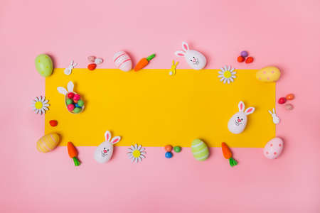 Top view festive Easter pink background with yellow frame made of colored eggs, decorative carrots,...