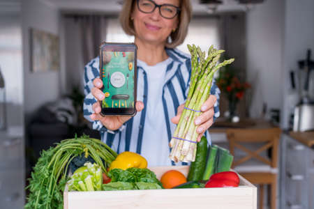 Woman holding a phone with active online mobile app of Healthy diet program and fresh asparagus bunch. New Start of healthy lifestyle, weight loss. Healthy food delivery, recipe box. Selective focus.の写真素材