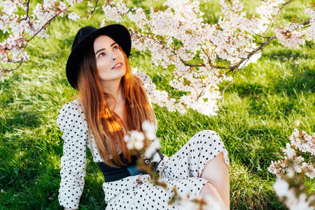 Young brunette woman in hat and dress sitting near white pink blooming tree. Generation Z girl enjoy...