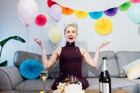 Portrait of a happy excited woman with birthday hat sitting on the couch and throwing confetti....